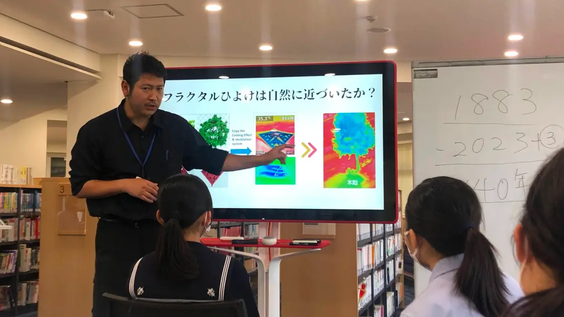 図書館のような空間で、スライドを使って授業を行う男性教員と生徒たちの様子。